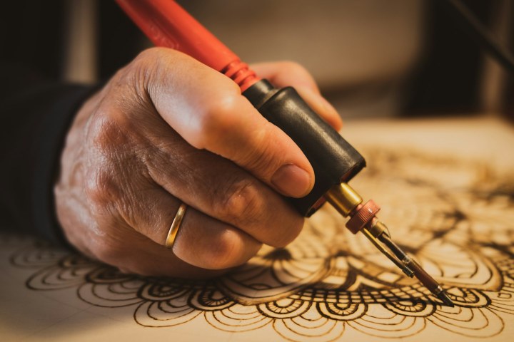 Kırmızı saplı pirografi kalemiyle açık renkli yüzeye çizgilerle mandala yakılan el